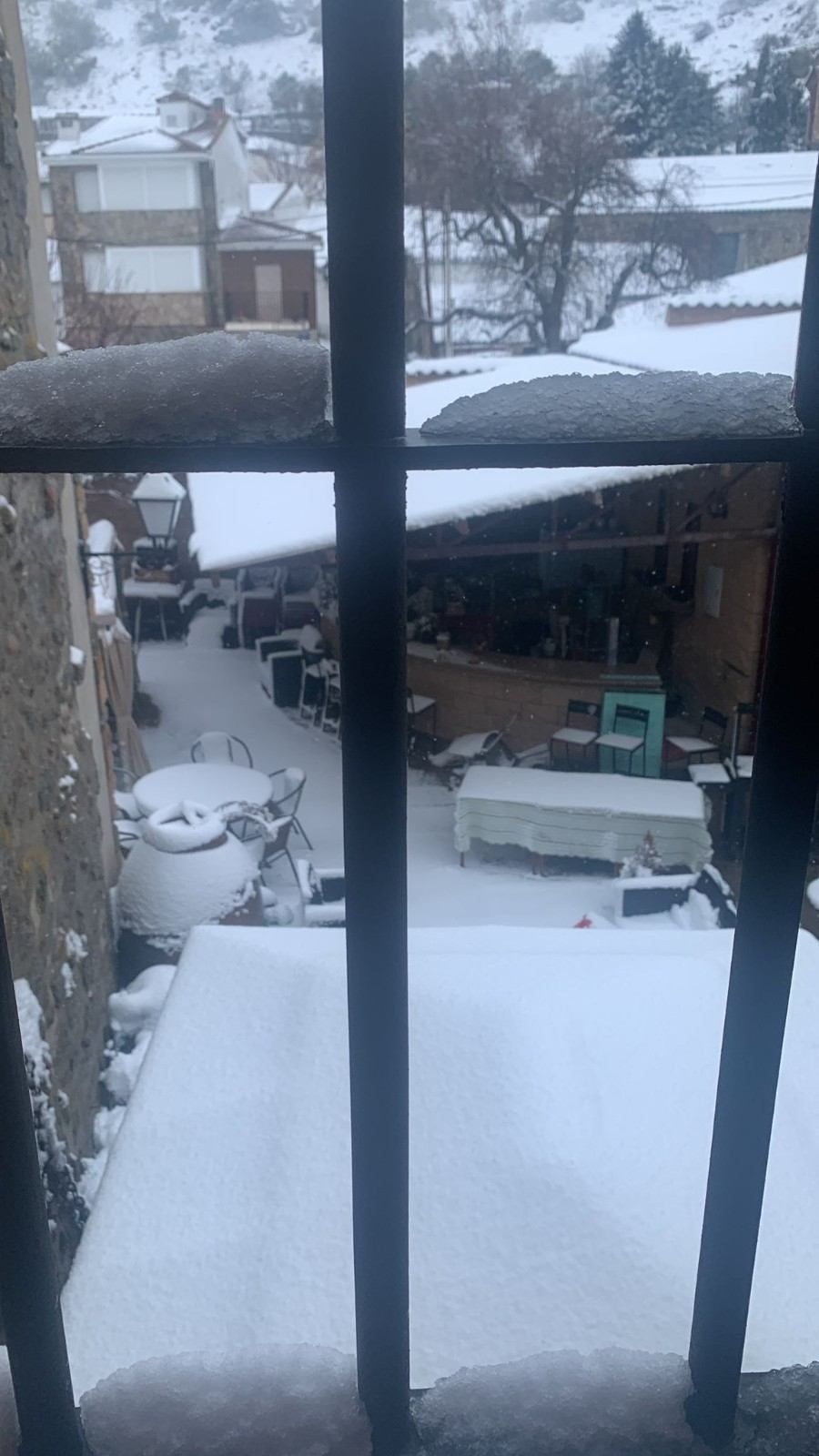 Patio y tejados cubiertos de nieve vistos a través de una ventana, con un coche aparcado y árboles al fondo.