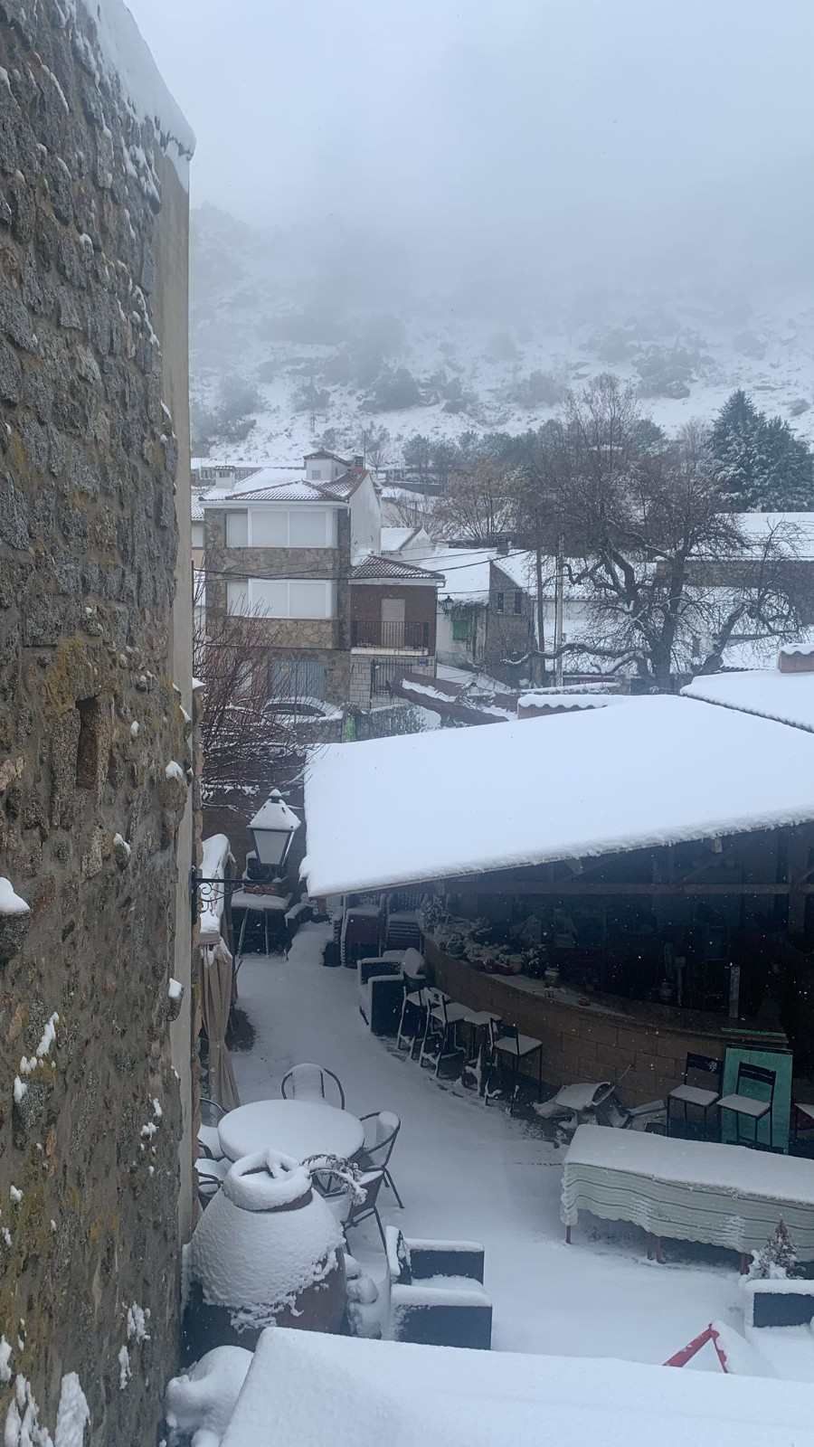 Patio nevado con edificios, árboles y una ladera neblinosa al fondo.