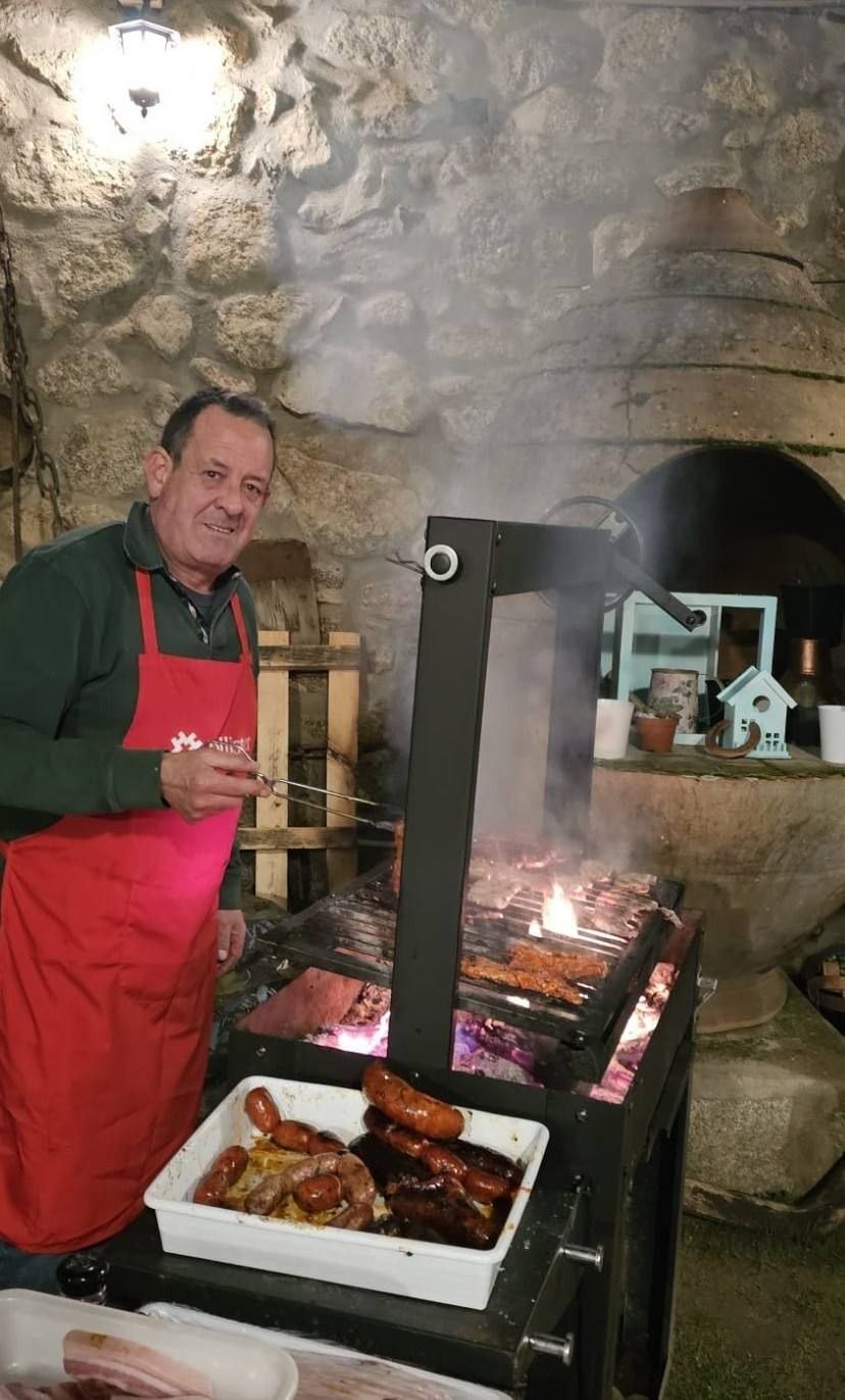 Un hombre asa carne junto a una chimenea de piedra, con un delantal rojo, sonriendo a la cámara.