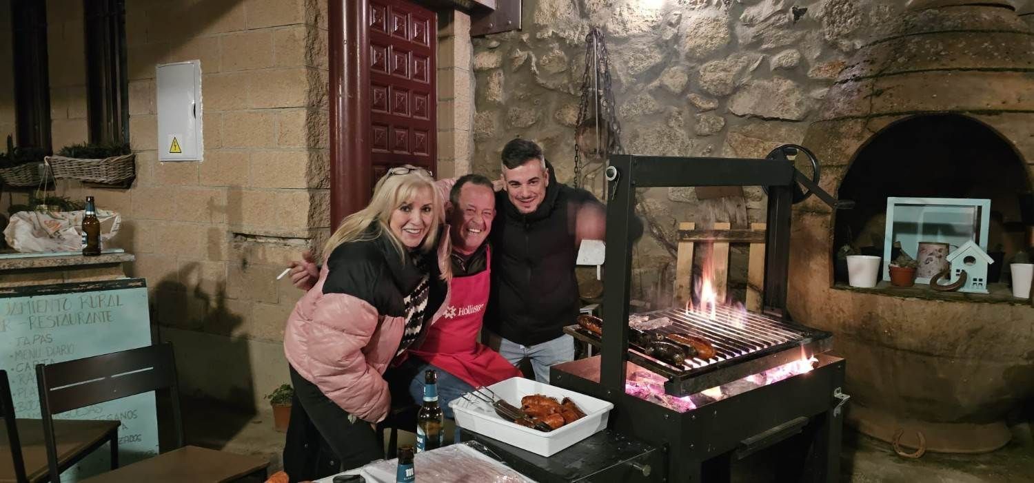 Tres personas sonriendo junto a una chimenea encendida en una habitación rústica de piedra, con bandejas de comida frente a ellas.