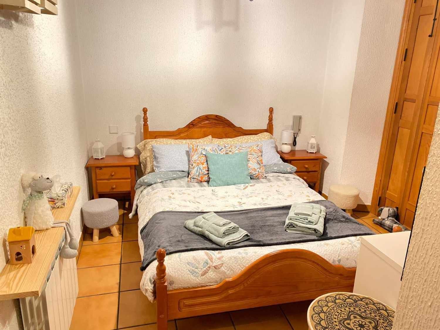 Habitación pequeña con cama de madera, ropa de cama gris, almohada azul y mesitas de noche sobre suelo de baldosas.
