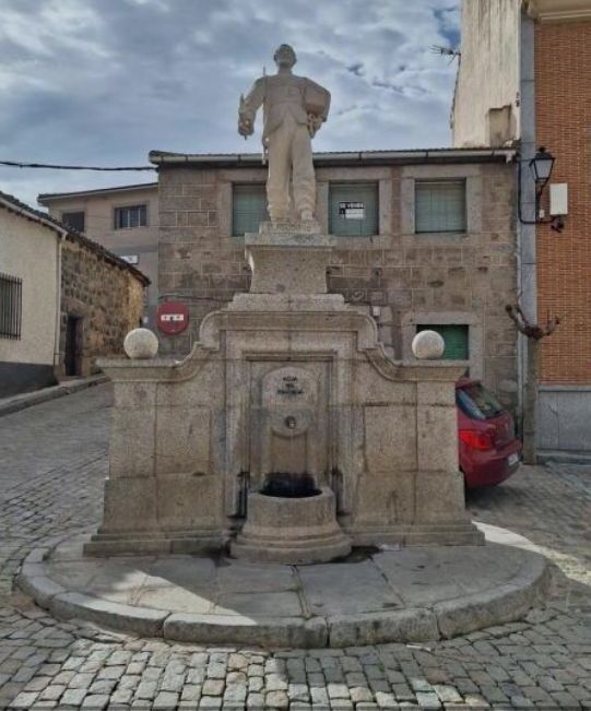 Fuente de piedra con una estatua de pie en una plaza de pueblo empedrada, junto a edificios antiguos y un coche rojo aparcado.