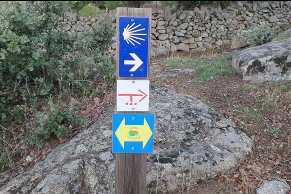 Señalización de sendero con flechas azules, blancas y amarillas junto a un camino rocoso y un muro de piedra.