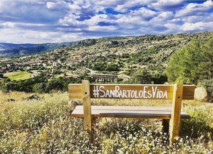 Banco de madera con letrero “#SanBartoloEsVida” con vistas a un paisaje verde en ladera bajo cielos nublados.