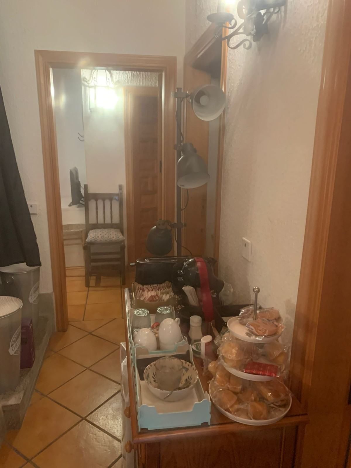 Pasillo estrecho con suelo de baldosas de terracota, una silla al final y una mesa auxiliar repleta de platos y utensilios de cocina.