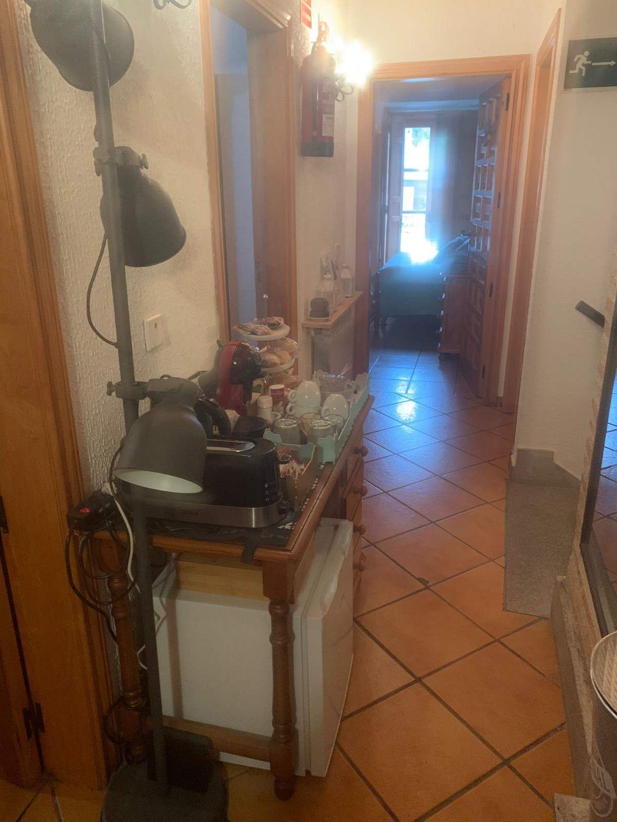 Pasillo estrecho con suelo de baldosas, una mesita, objetos colgados y una puerta luminosa al final.
