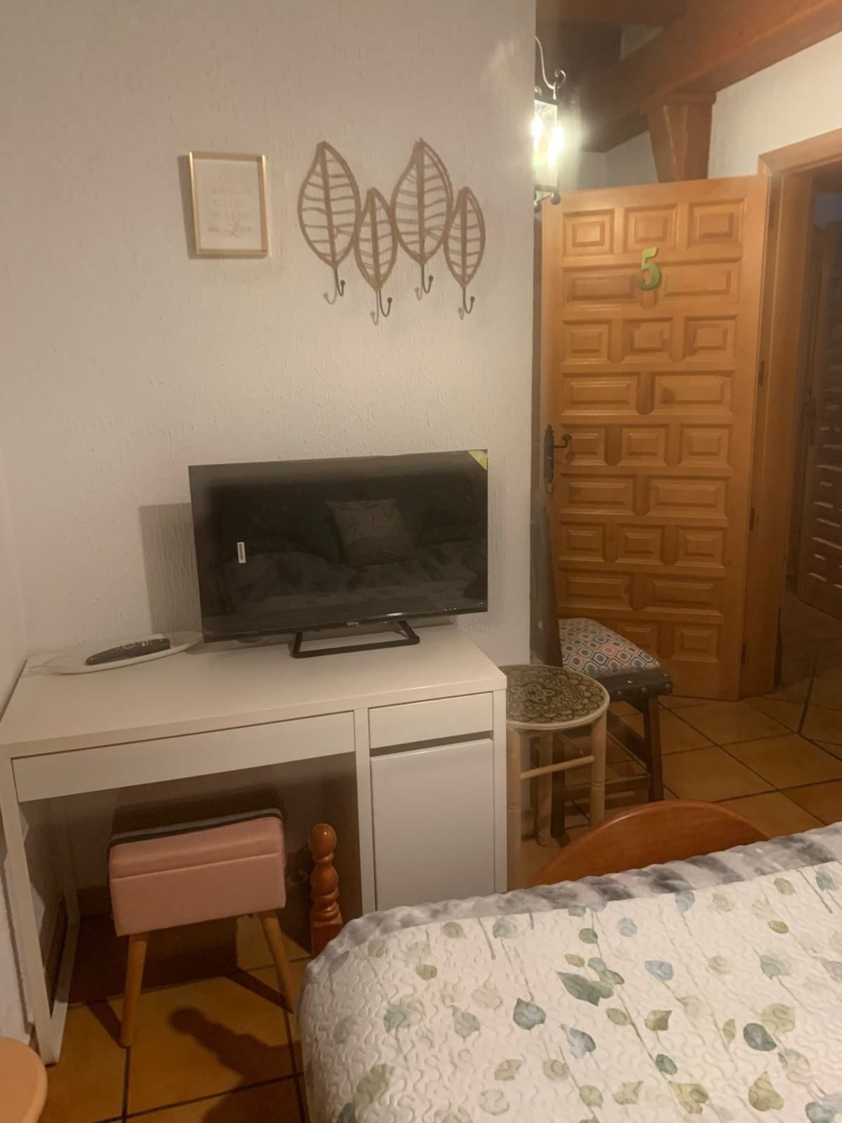 Habitación pequeña con escritorio y televisor, puerta de madera, decoración en la pared y una cama en primer plano.