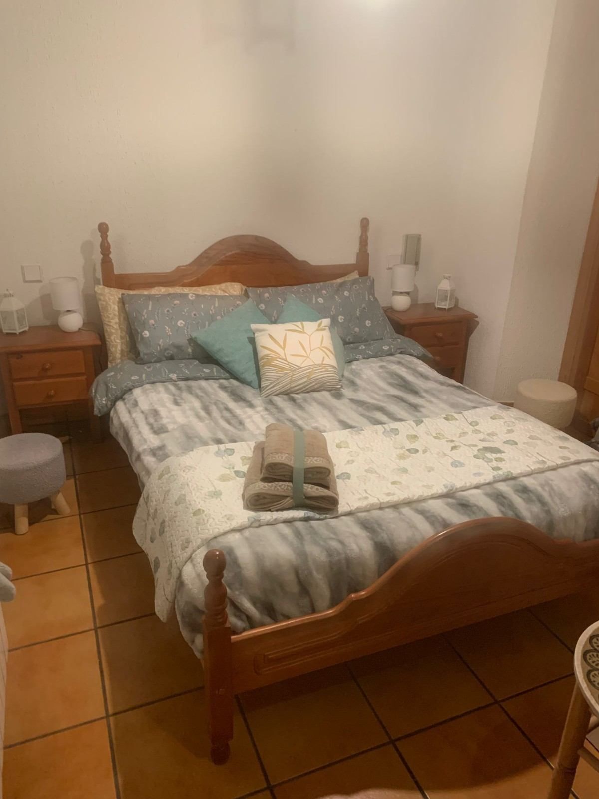 Cama de madera con ropa de cama gris y almohadas color turquesa en un pequeño dormitorio con suelo de baldosas, flanqueada por mesitas de noche a juego.