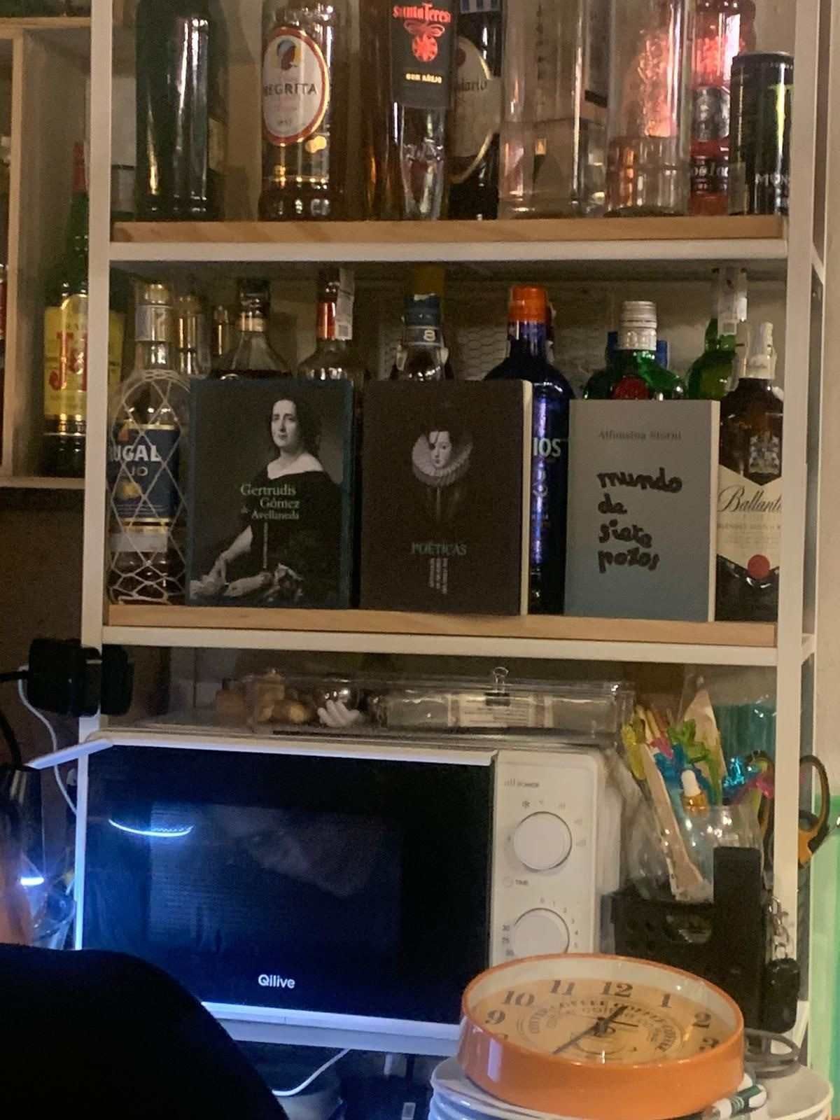 Estantería de madera con botellas de licor sobre un escritorio con un ordenador portátil y un reloj naranja en una habitación con poca luz.