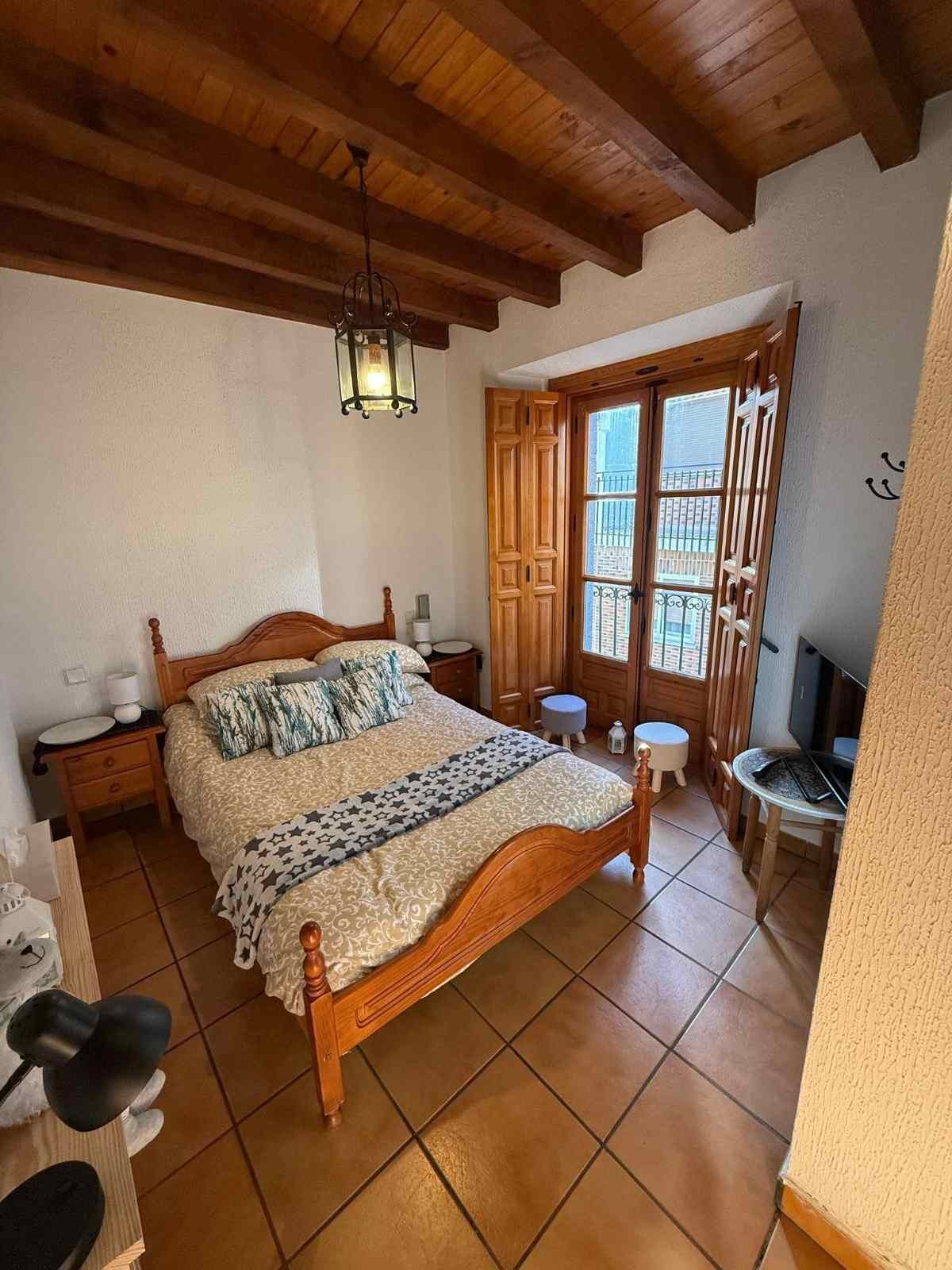 Habitación rústica con techo de madera, suelo de baldosas, cama de madera y puertas que dan al balcón y dejan entrar la luz.