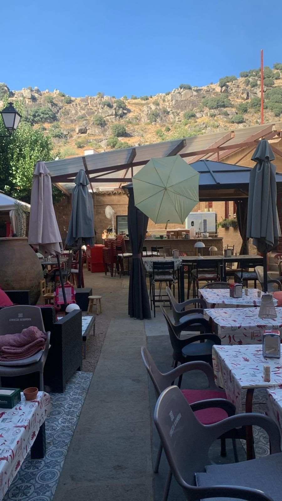 Terraza de un restaurante al aire libre con mesas y sombrillas vacías junto a una ladera rocosa bajo un cielo azul.
