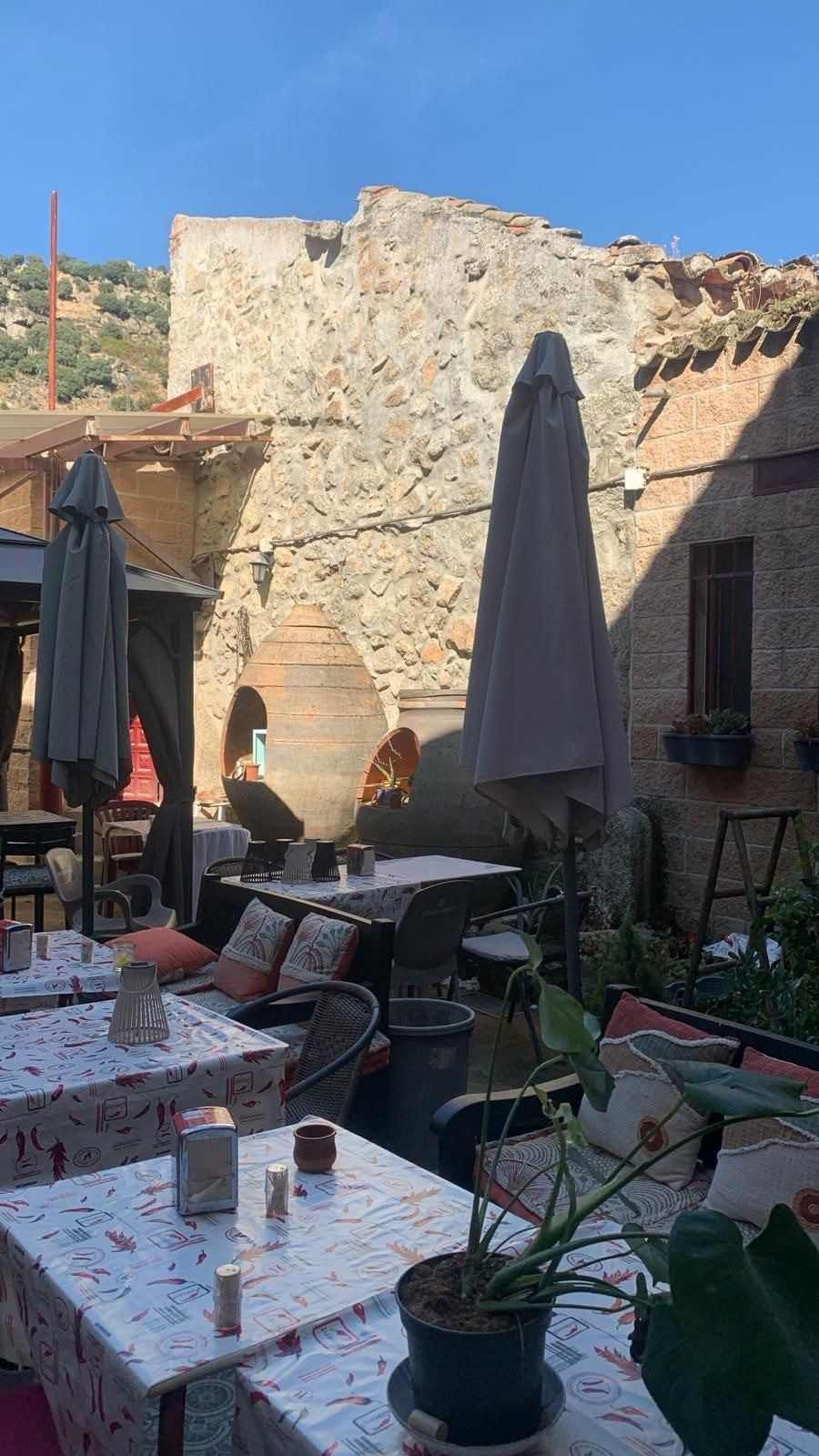 Terraza de una cafetería al aire libre con mesas vacías, sombrillas, macetas y un muro de piedra bañado por el sol.
