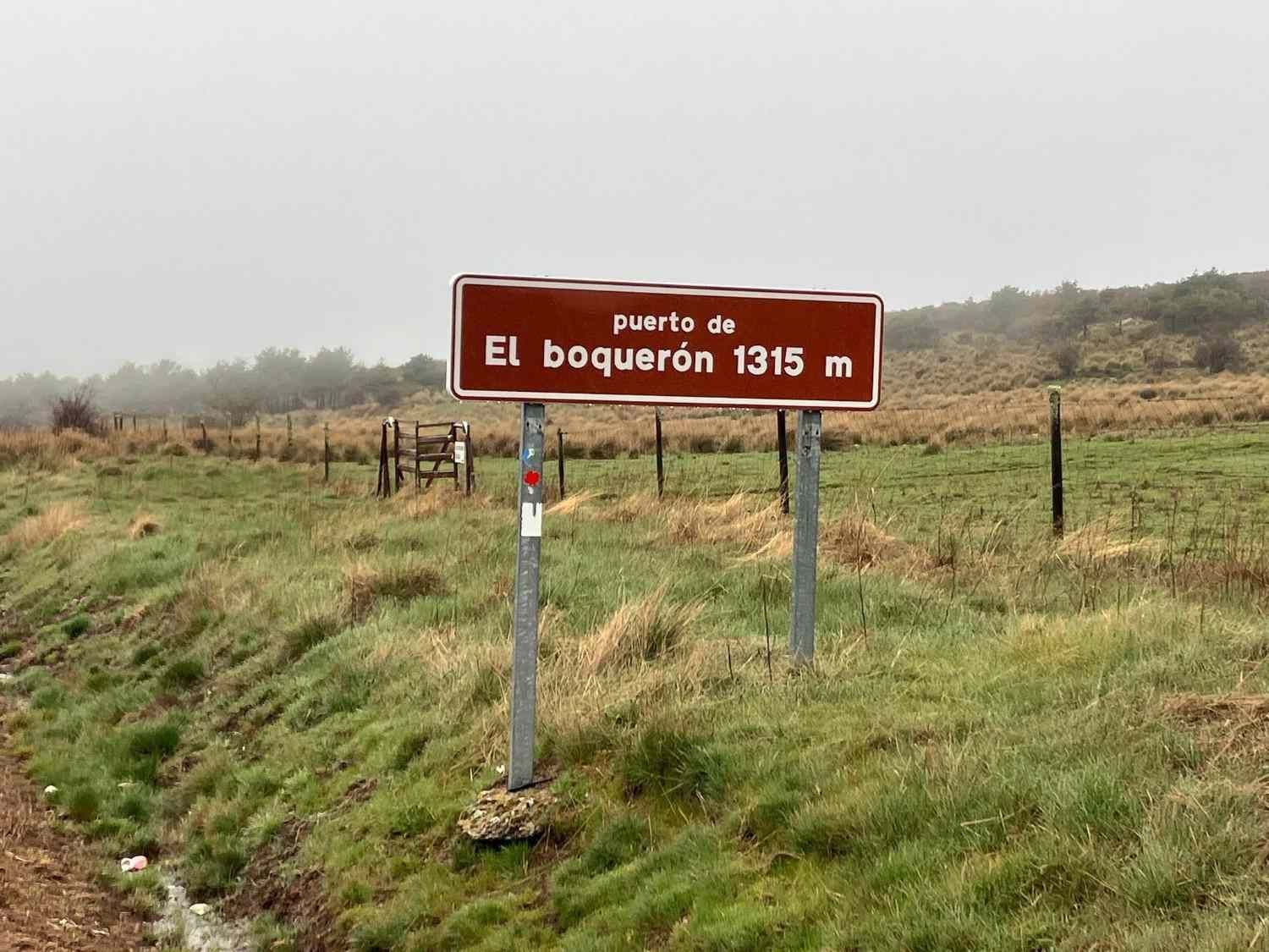 Un cartel marrón al borde de la carretera que dice “Biens du Boquerón 1315 m” en un campo de hierba bajo un cielo nublado.