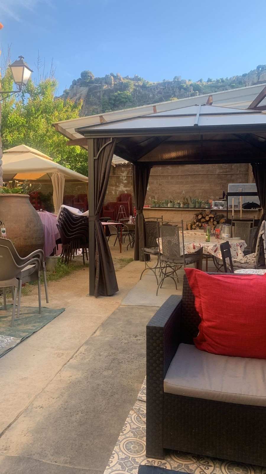 Terraza con asientos de mimbre, cojines rojos y un cenador con sombra con vistas a una ladera.