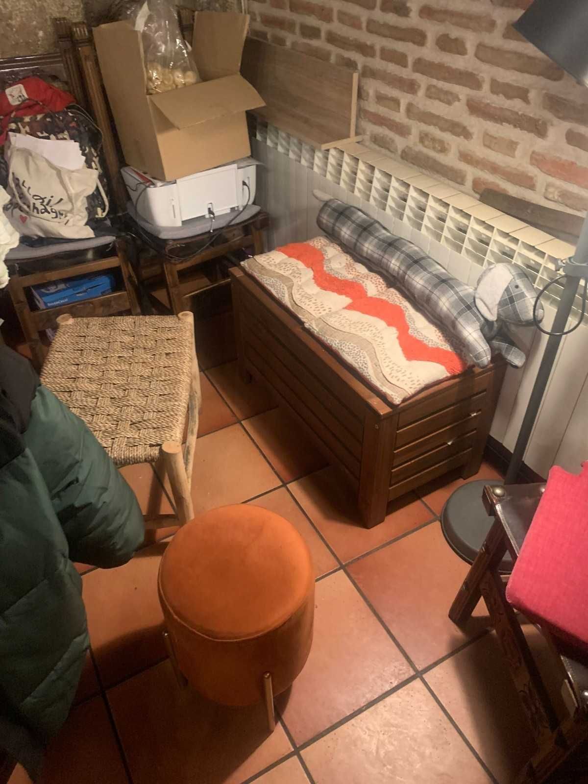 Habitación pequeña con paredes de ladrillo, un baúl de madera, un taburete naranja, cajas y un banco acolchado debajo de un ventilador de ventana.