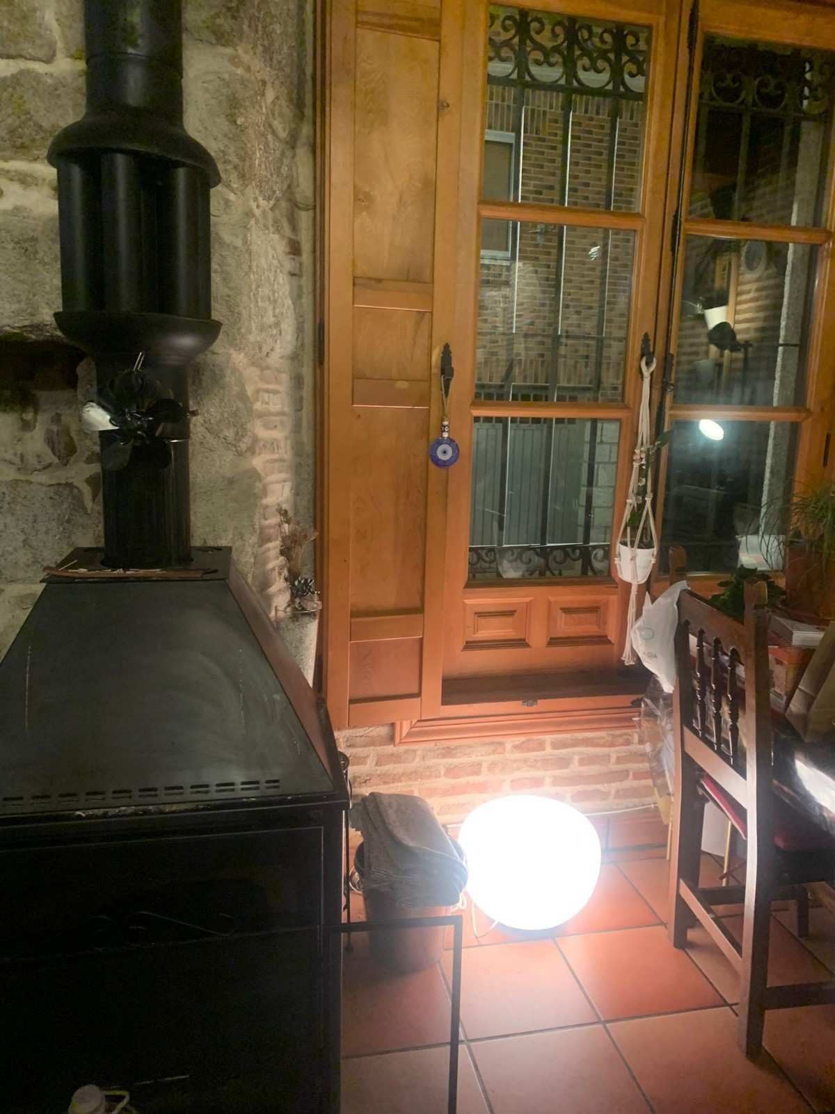 Interior acogedor con una estufa negra, puerta de madera, silla y una lámpara de pie encendida junto a la ventana.
