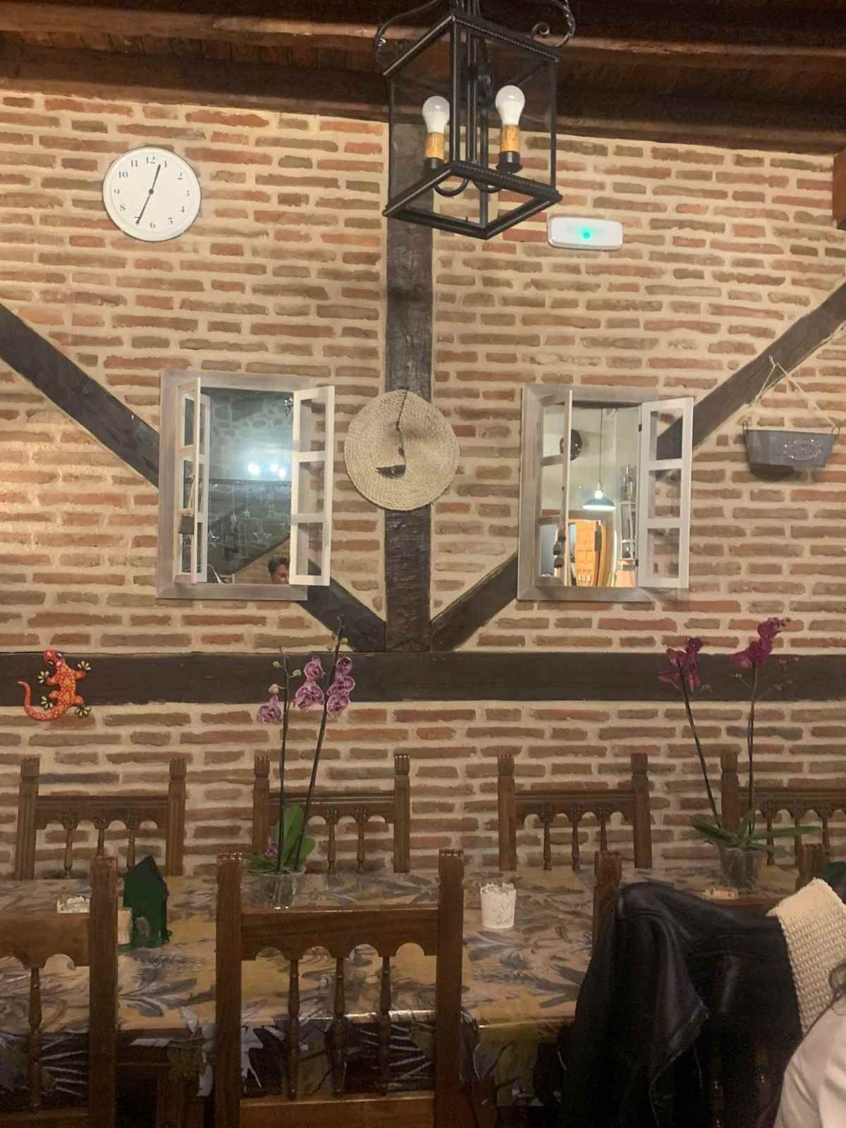 Interior de una cafetería de ladrillo con faroles colgantes, reloj de pared, ventanas abiertas y mesas dispuestas bajo una decoración rústica.