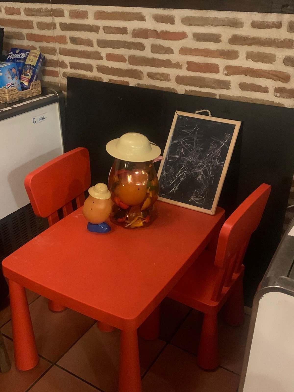 Mesita y sillas pequeñas de color rojo, del tamaño de un niño, con una lámpara y una pizarra en una esquina, contra una pared de ladrillos.