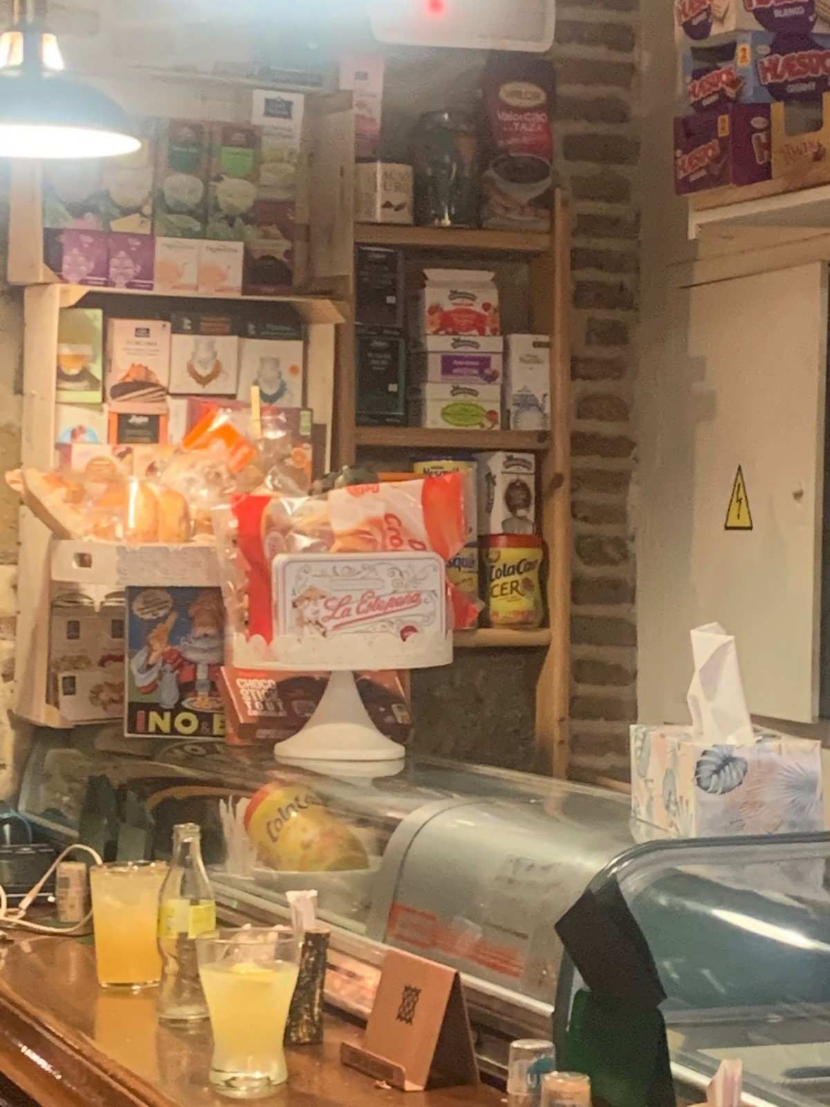 Acogedora barra de cafetería con pasteles, bebidas y estantes con aperitivos y decoración detrás de la caja registradora.