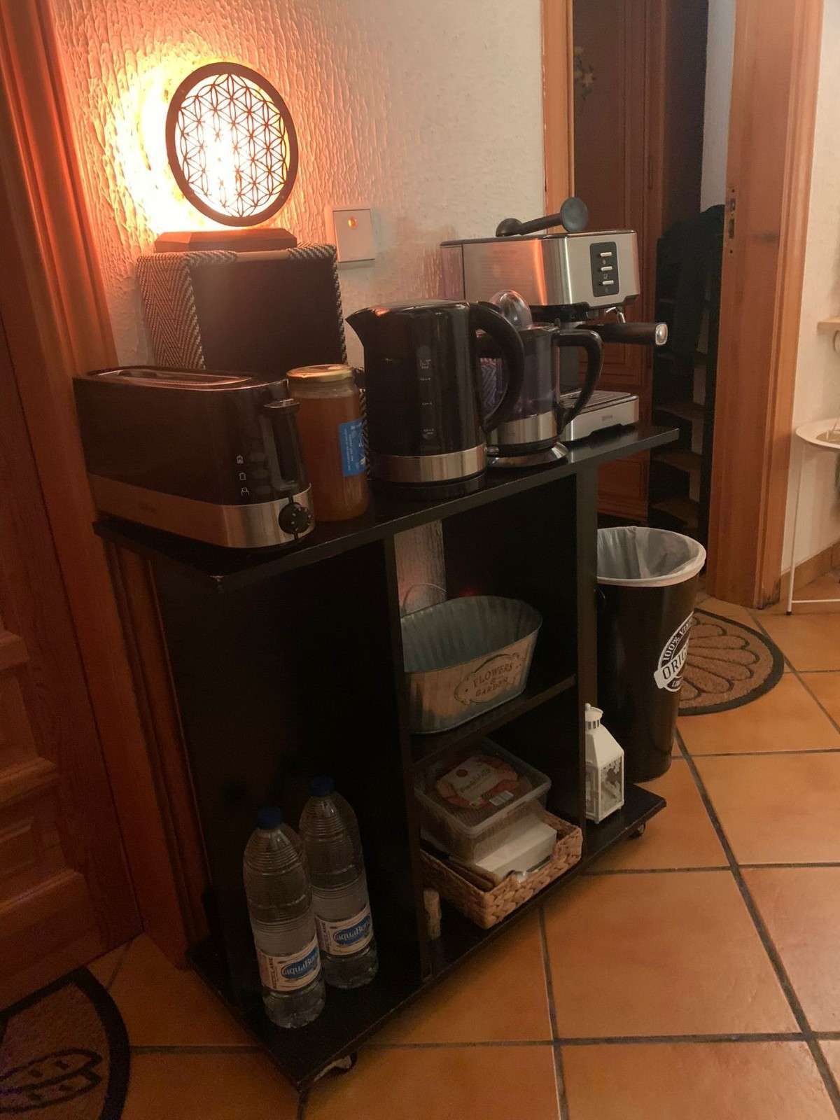 Estación de café con tazas, hervidor eléctrico y suministros en un estante negro en un rincón de la cocina alicatado.