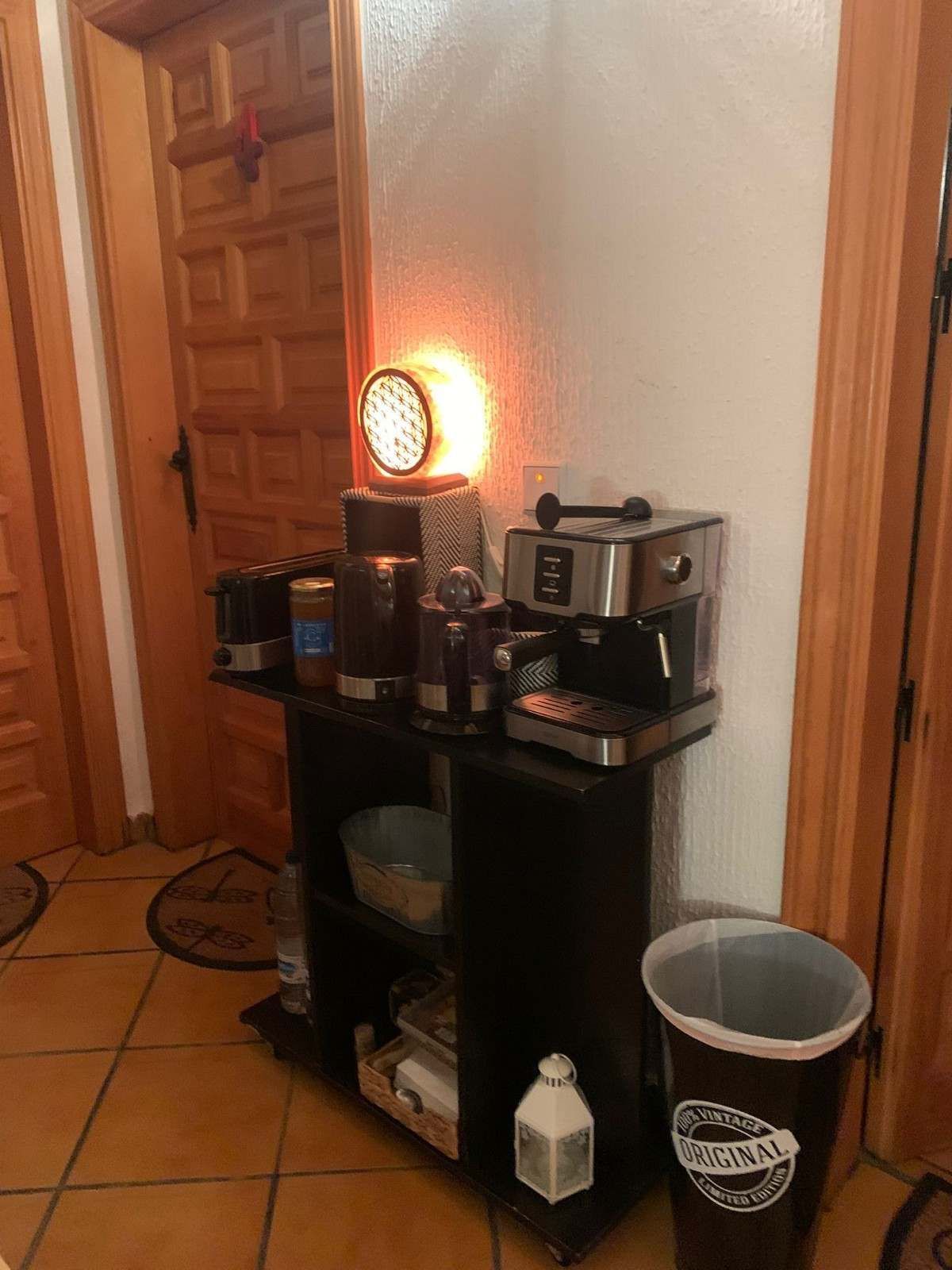 Pasillo con una estantería negra que contiene utensilios para café, una lámpara encendida y un cubo de basura junto a una puerta de madera.