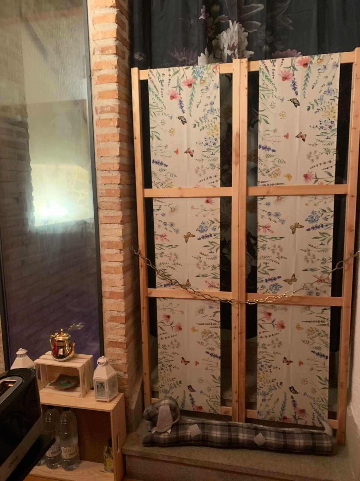 Pequeño banco junto a la ventana con paneles florales, marco de madera y cojín a cuadros, situado junto a una pared de ladrillos y una ventana oscura.