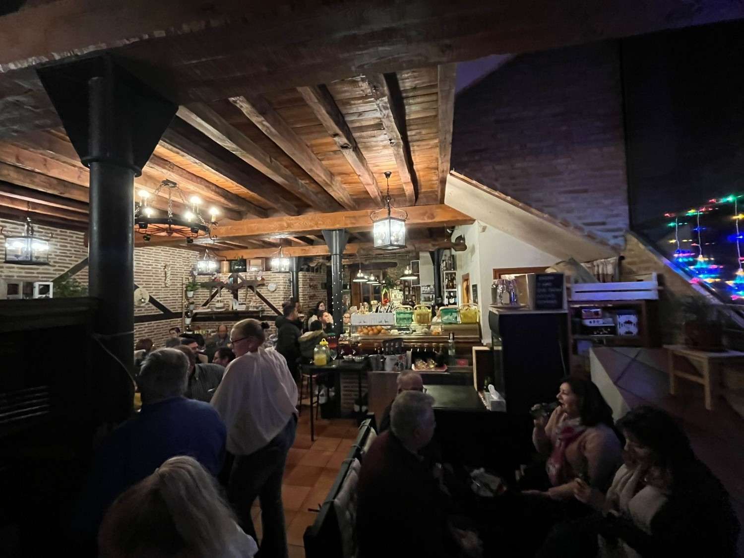 Interior de bar rústico con gente sentada en mesas bajo luces cálidas y vigas de madera a la vista.