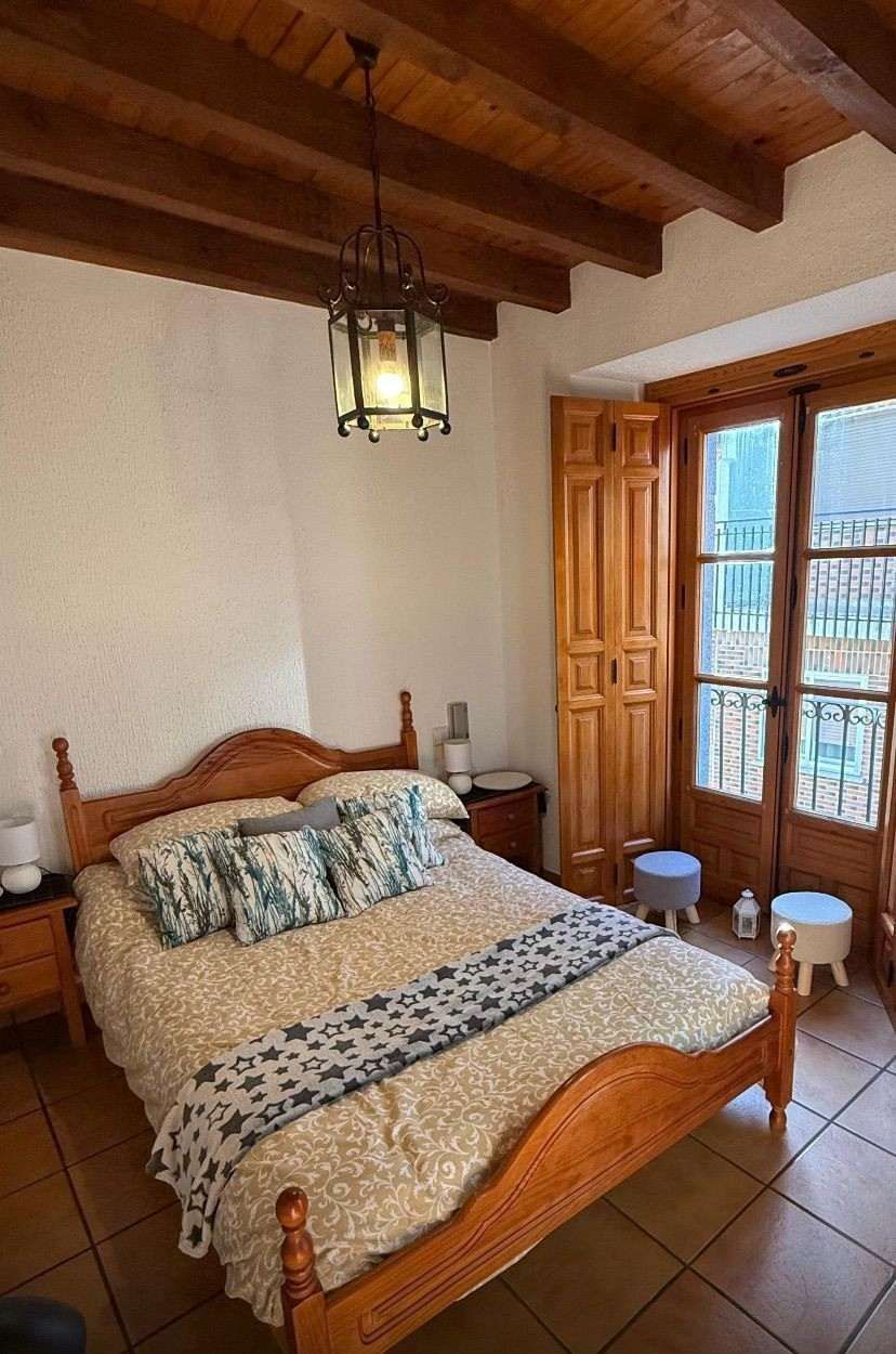 Habitación acogedora con cama de madera, suelo de baldosas, vigas vistas y ventana con puertas abiertas.