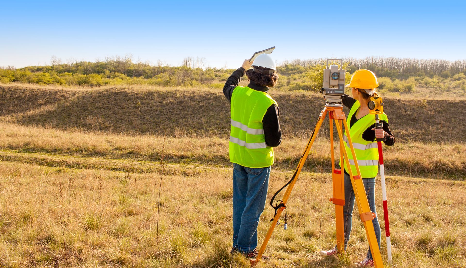 Dos topógrafos con chalecos de seguridad utilizando equipos de topografía al aire libre en un campo.