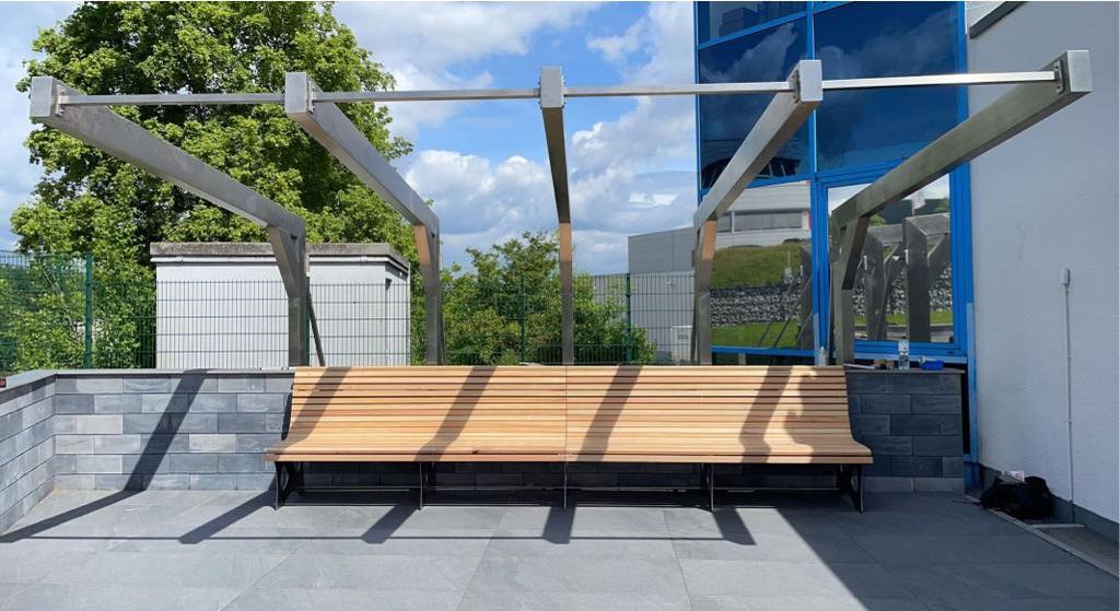 Holzbank unter einer Metallpergola auf einem Dach, mit einem blau getönten Fenster im Hintergrund.