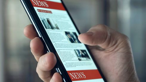 Person mit Smartphone in der Hand, die in einem Gebäude durch eine Nachrichten-Website mit rotem Banner scrollt.