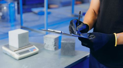 Person mit Handschuhen, die in einem blau beleuchteten Labor neben Betonwürfeln und einer digitalen Waage auf einem Klemmbrett schreibt.
