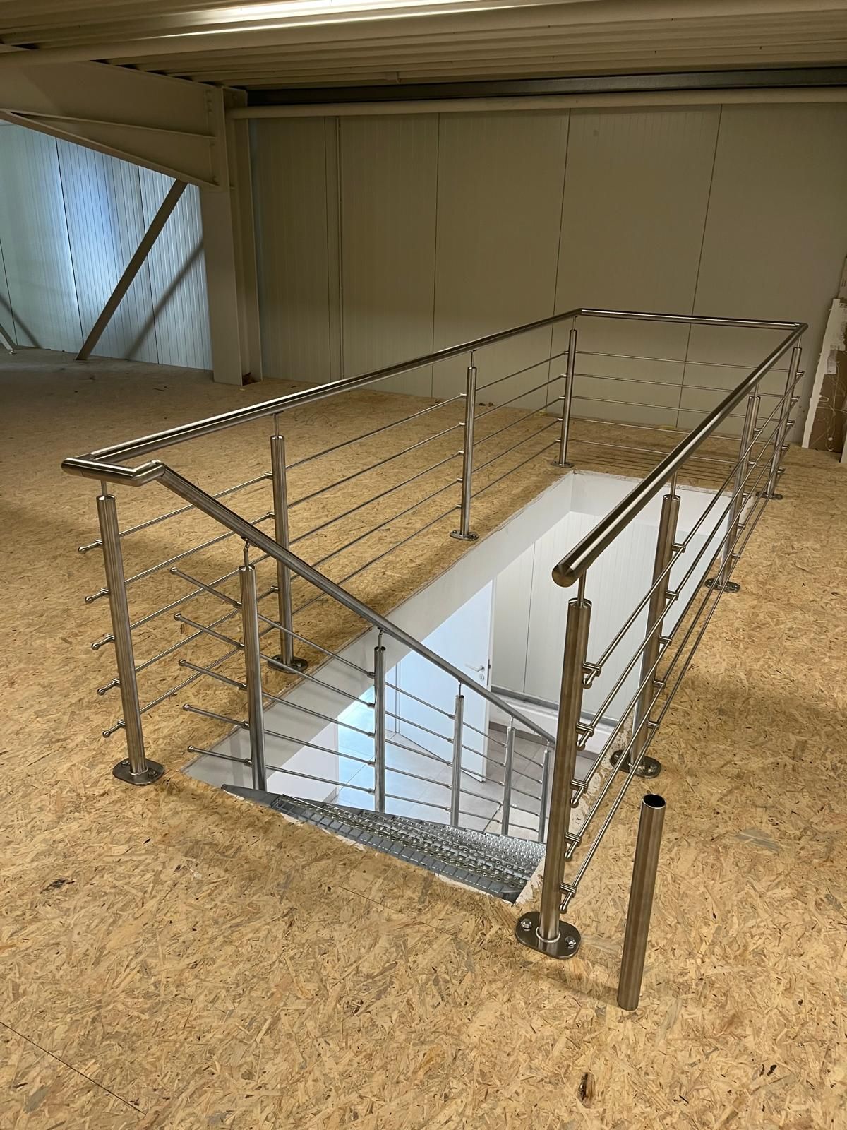 Handläufe aus Edelstahl umgeben eine Treppe, die in einen Bereich mit weißen Wänden hinabführt; Boden und Wände aus Sperrholz befinden sich in einem großen Raum.