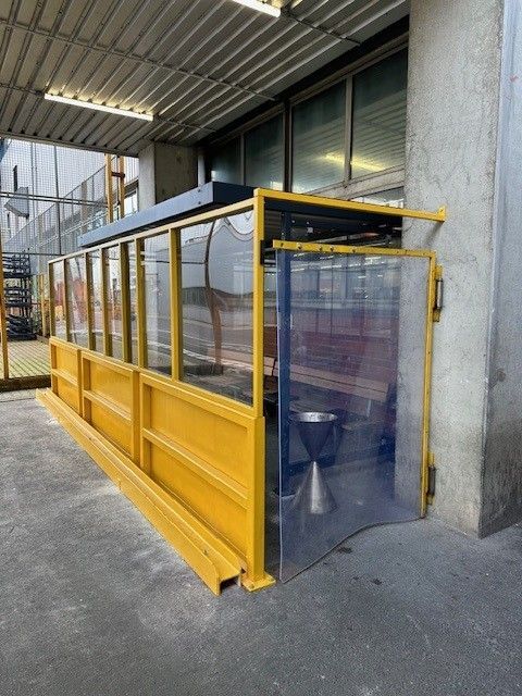 Gelber und durchsichtiger, durch eine Barriere abgegrenzter Sitzbereich im Freien mit einem einzelnen Metallmülleimer.