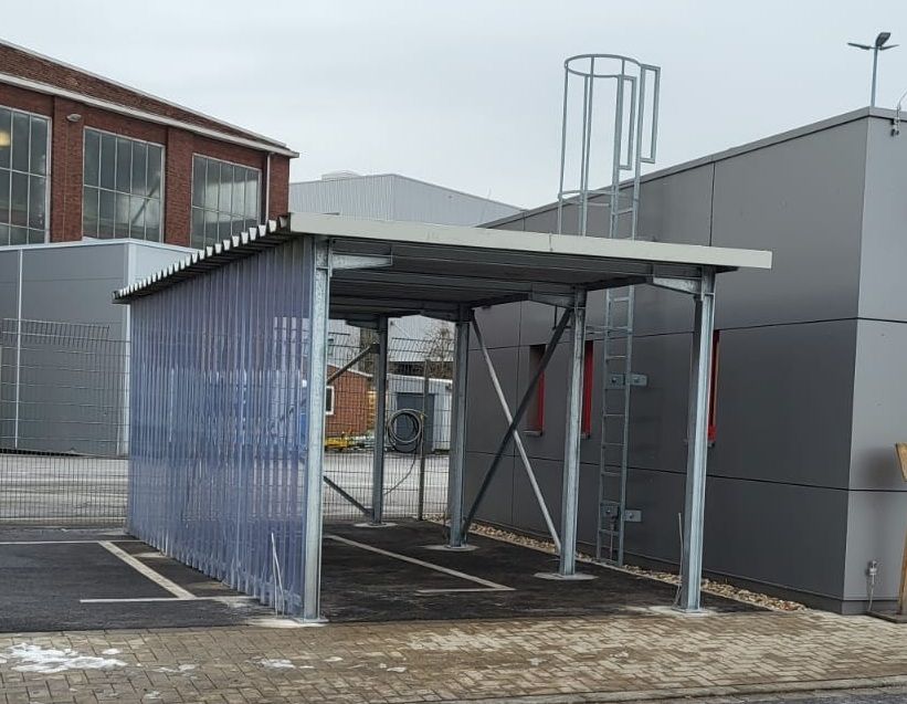 Fahrradunterstand aus Metall mit durchscheinenden Seitenwänden und einer Leiter vor einem grauen Gebäude.