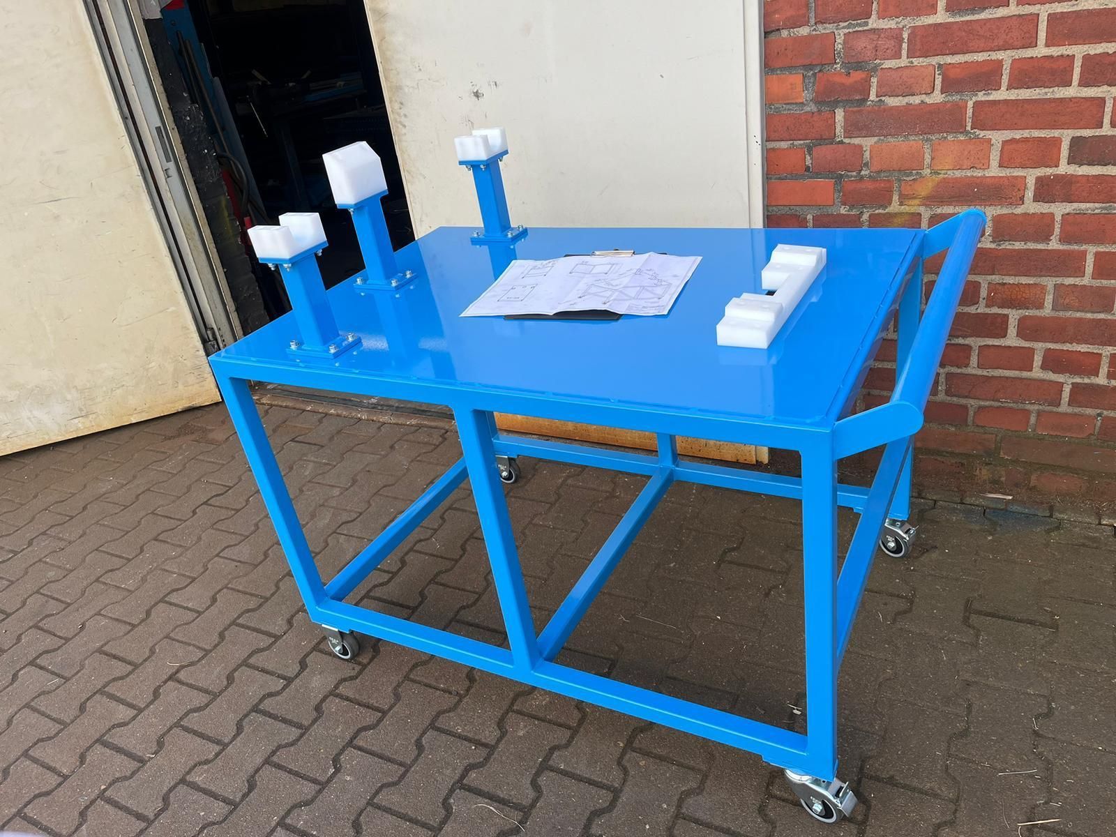 Blauer Metallwagen mit Tisch, Rädern und Papier darauf, neben einer Ziegelmauer.
