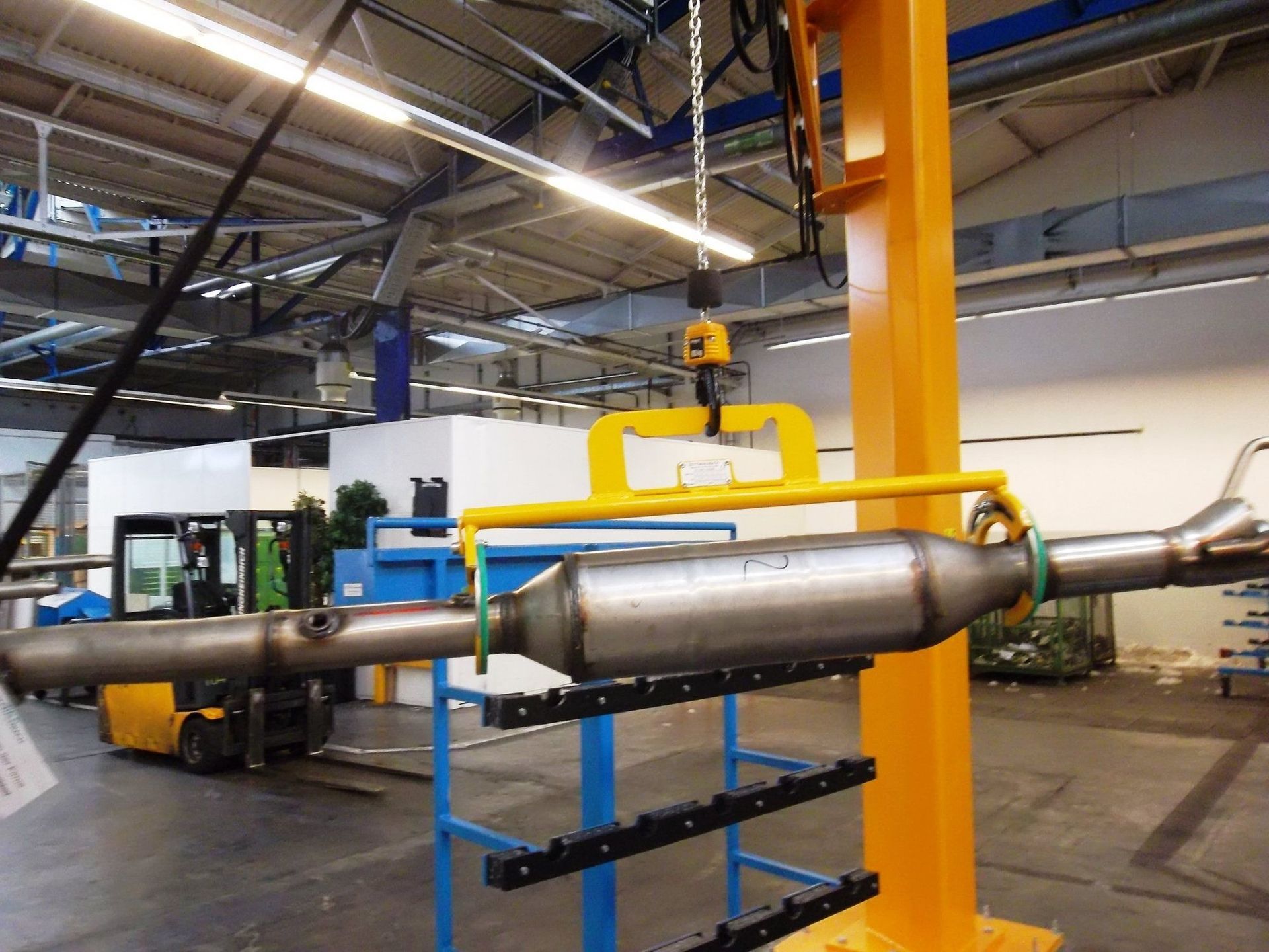 Ein gelber Flaschenzug hebt in einer Fabrik ein Autoauspuffteil an. Im Hintergrund ist ein Gabelstapler zu sehen.