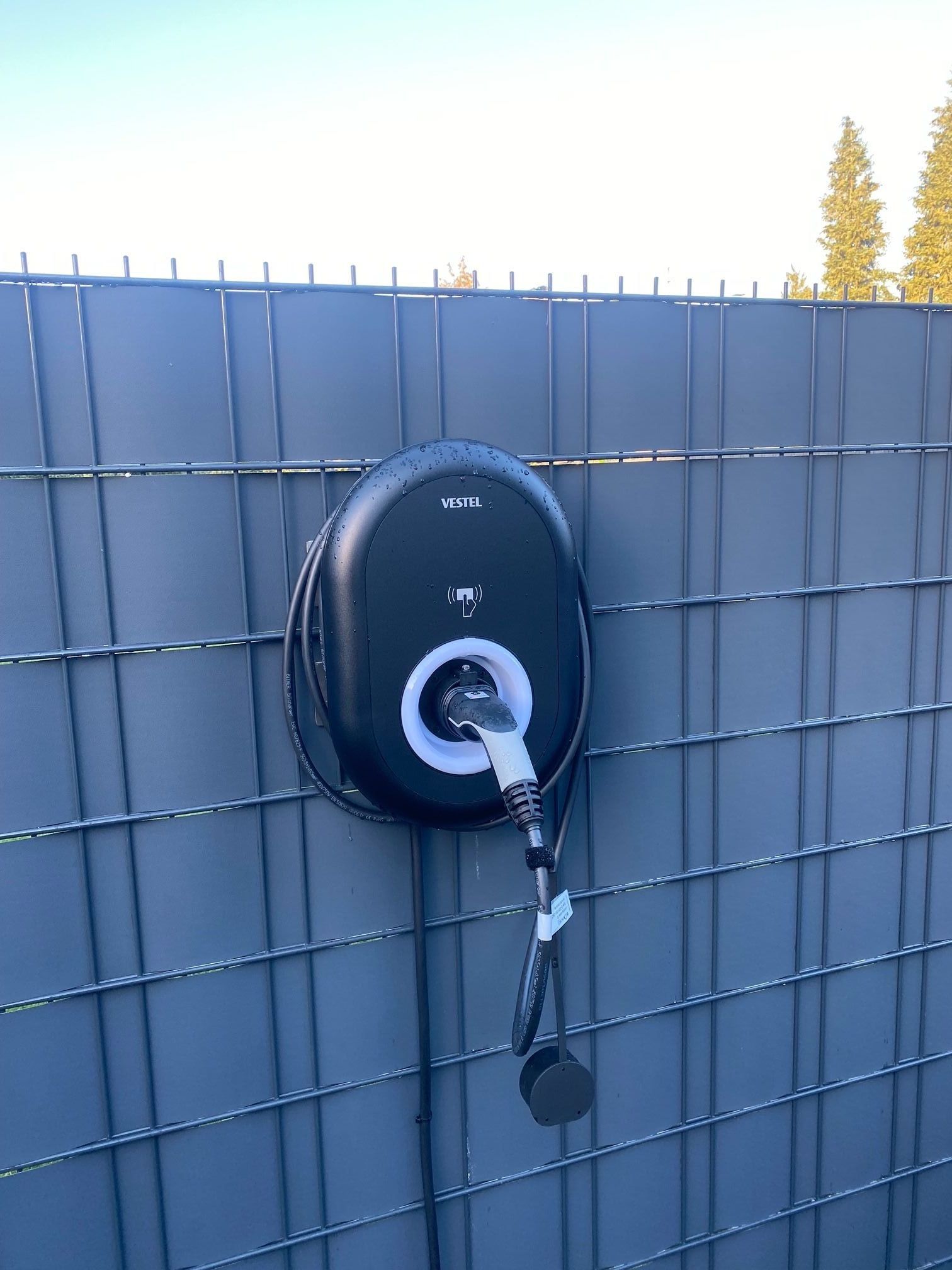 Schwarzes EV-Ladegerät, montiert an einem grauen Zaun, mit eingestecktem Ladekabel.