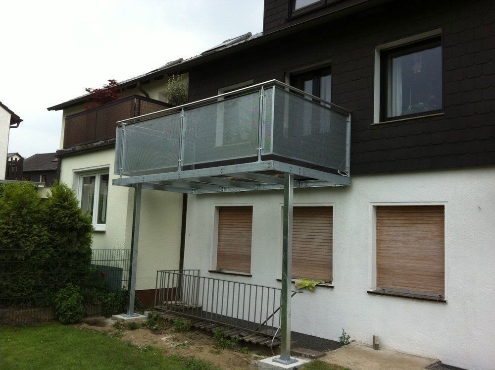Metallbalkon mit Glasgeländer, gestützt durch Pfosten, an einem zweistöckigen Gebäude.