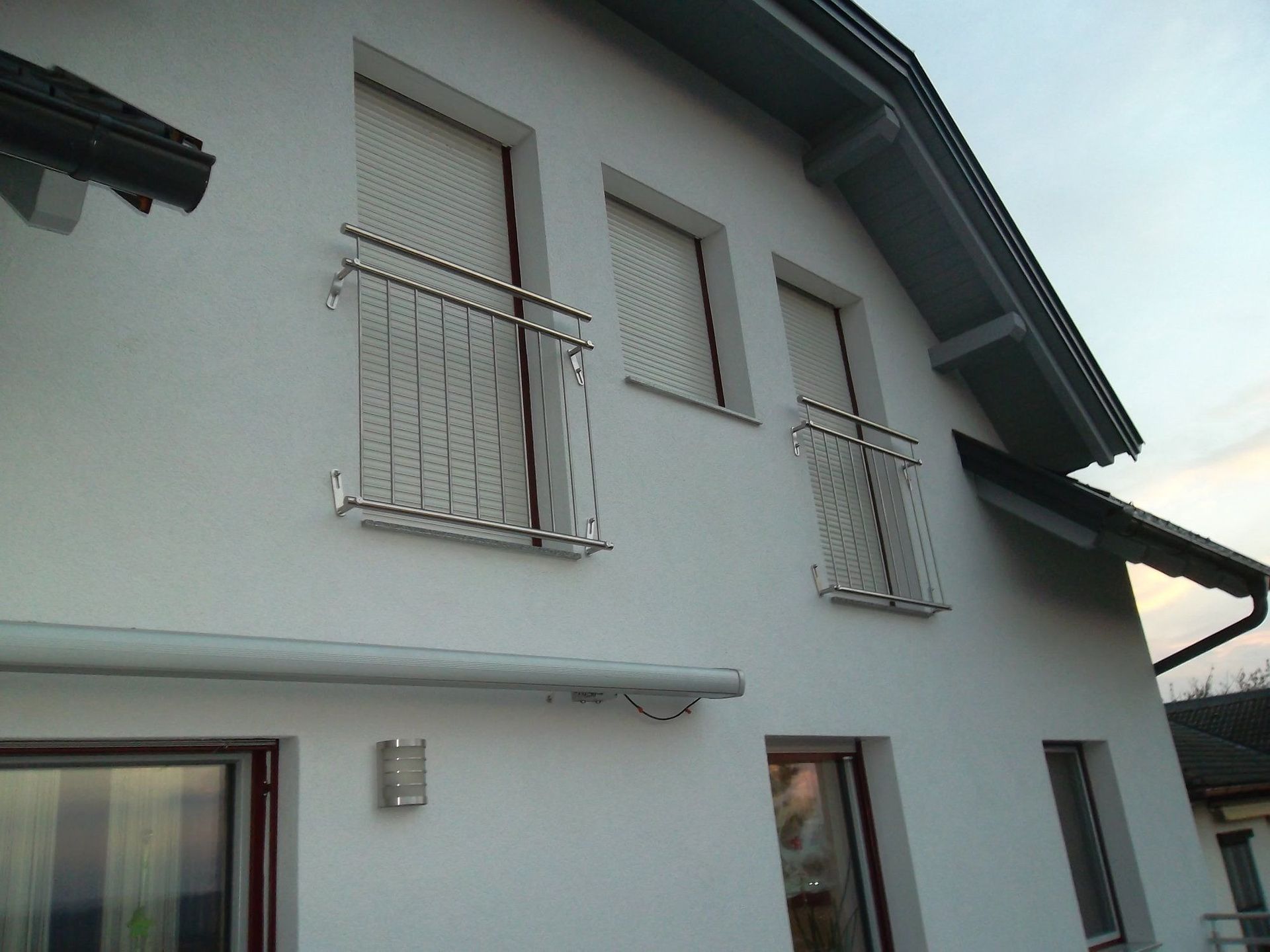 Weißes Hausäußeres mit zwei kleinen Balkonen. Silbernes Geländer, Fenster und graues Dach.