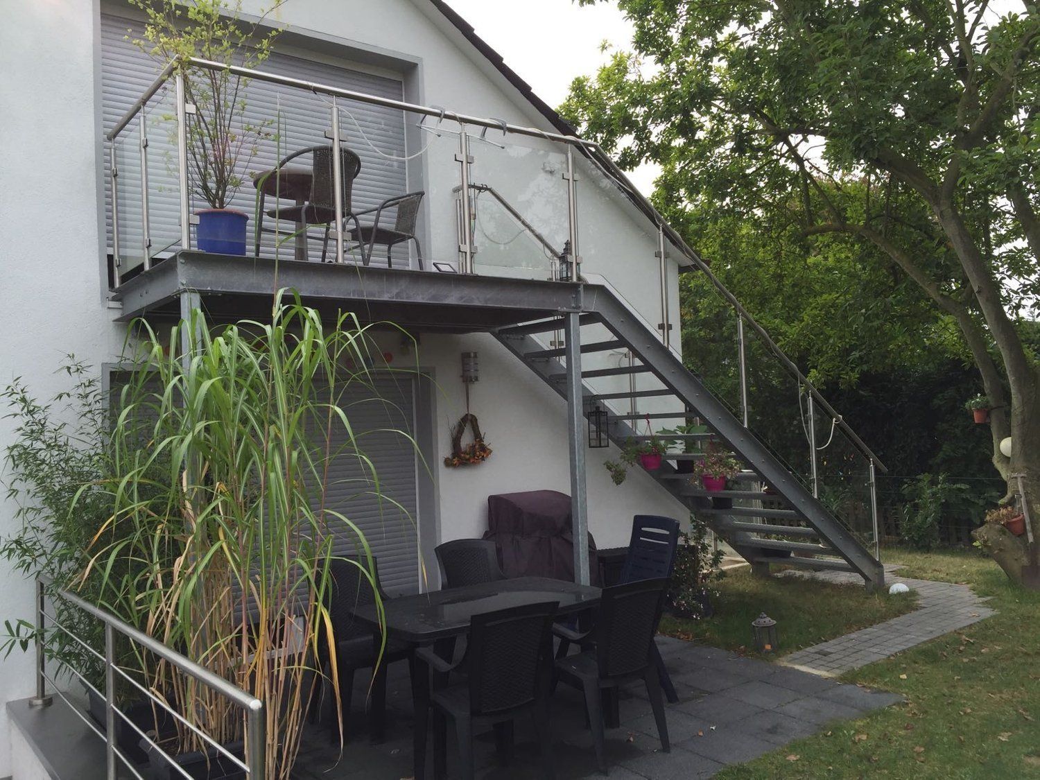 Balkon und Außentreppe aus Metall auf einem weißen Haus. Gartenmöbel auf der Terrasse. Grünes Gras und Bäume.