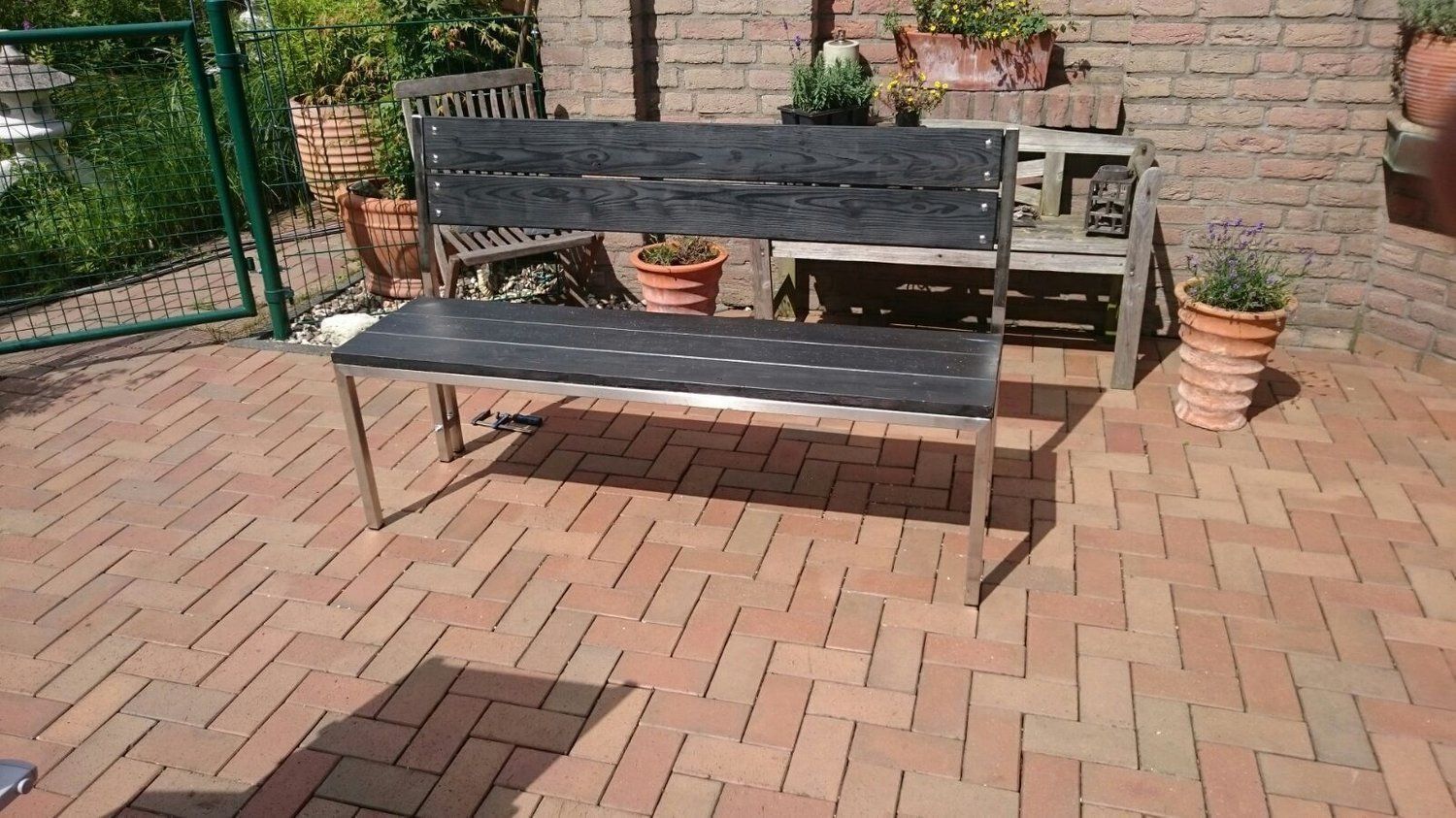 Eine schwarze Bank auf einer gemauerten Terrasse mit Pflanzen im Hintergrund; sonnige Szene im Freien.