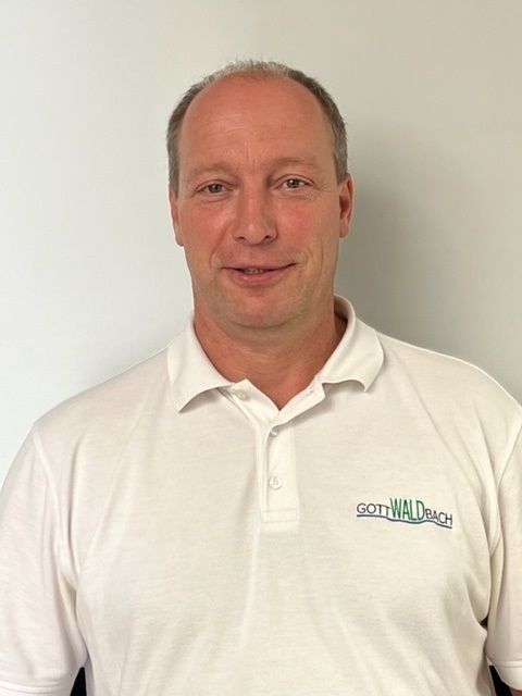 Ein Mann mittleren Alters mit heller Haut und weißem Poloshirt steht vor einer weißen Wand.