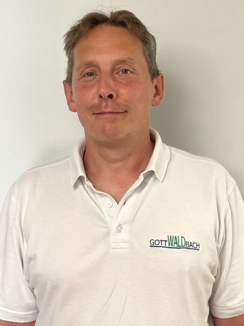 Mann mittleren Alters in einem weißen Poloshirt mit dem Logo „GOFF WALD BACH“, lächelnd, vor einem schlichten weißen Hintergrund in die Kamera blickend.