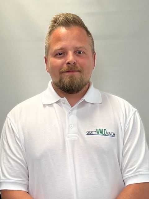 Mann mit Bart im weißen Poloshirt, Schriftzug „GOTTWALD WACH“, blickt in die Kamera.