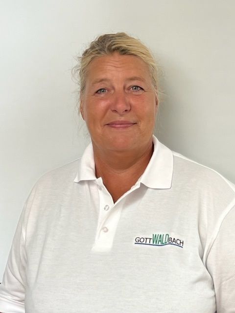 Blonde Frau im weißen Poloshirt mit grünem Text, leicht lächelnd. Weißer Wandhintergrund.