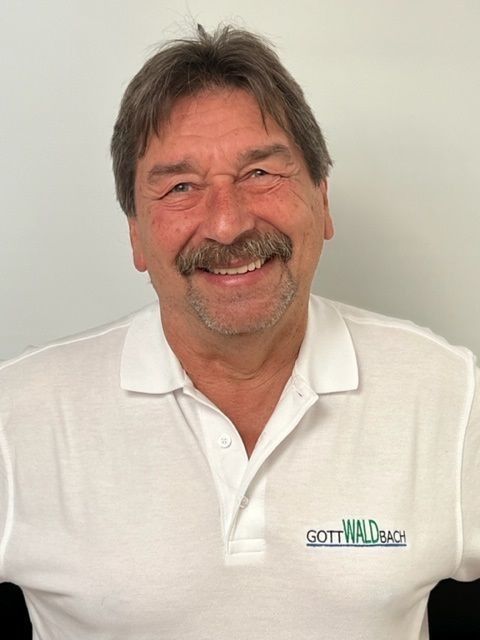 Lächelnder Mann mit Schnurrbart, der ein weißes Poloshirt mit der Aufschrift „GOTTWALD BACH“ trägt.