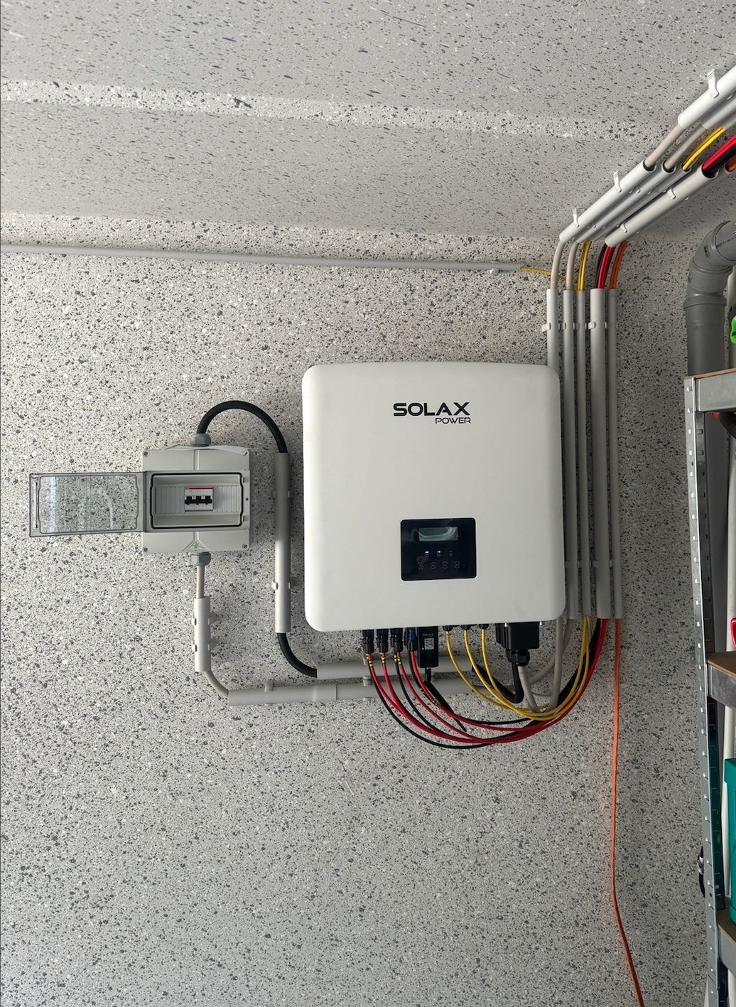 Weiße Solarwechselrichterbox an einer gesprenkelten Wand, verbunden mit Leitungen und Kabeln.