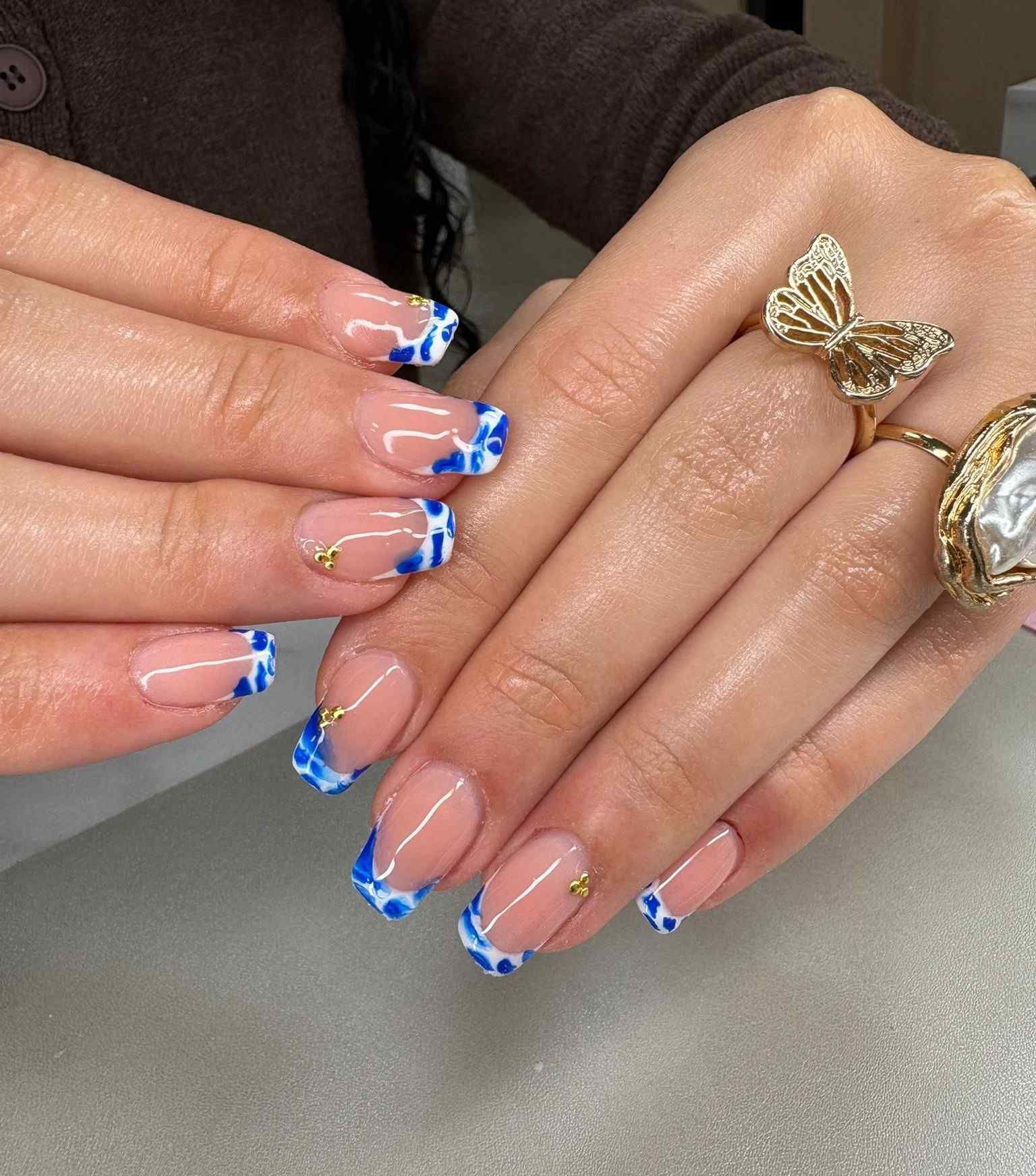 Manos con uñas pintadas con punta francesa en azul y blanco y anillos de oro descansando sobre una mesa.