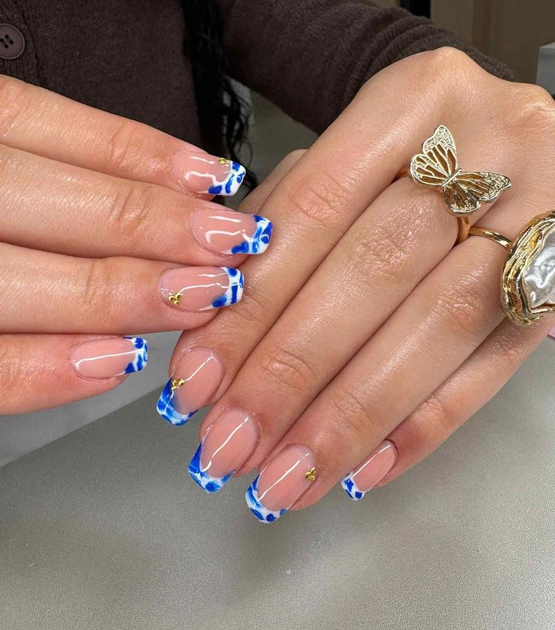 Manos con uñas pintadas con punta francesa en azul y blanco y anillos de oro descansando sobre una mesa.